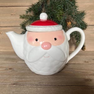 Puede incluir: Tetera de cerámica blanca con forma de Papá Noel, con tapa roja y mejillas rosas. La tetera tiene un asa y un pico vertedor, perfecta para servir té durante la temporada navideña.