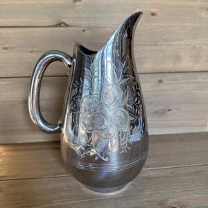 May include: A silver-colored pitcher with a curved handle and a spout. The pitcher is decorated with an etched floral design. The pitcher is likely made of metal and is suitable for serving beverages.