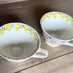 Op de afbeelding: Twee witte keramische theekopjes met gele bloempatronen en blauwe stippen langs de randen. De kopjes hebben handvatten met gele strepen en staan op een houten oppervlak. De binnenkant van de kopjes is wit.