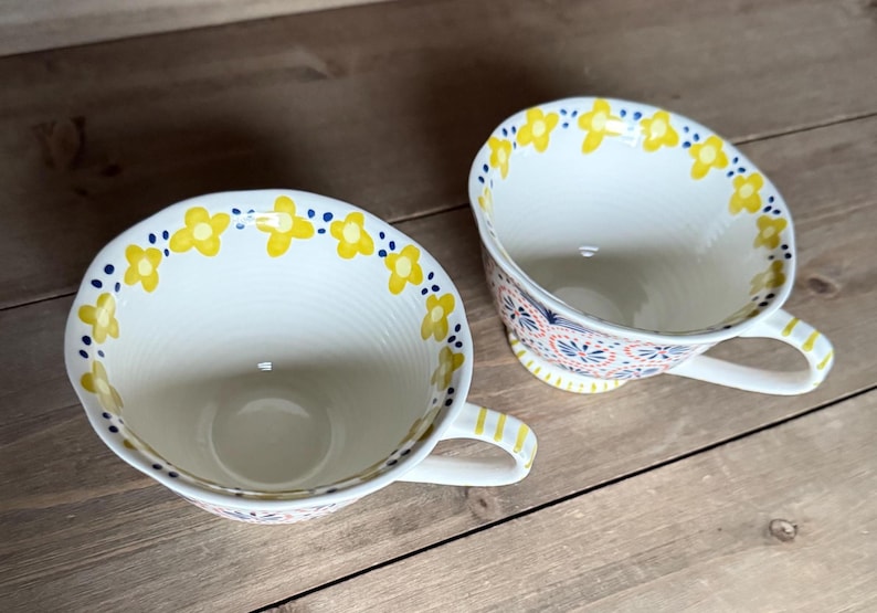 Op de afbeelding: Twee witte keramische theekopjes met gele bloempatronen en blauwe stippen langs de randen. De kopjes hebben handvatten met gele strepen en staan op een houten oppervlak. De binnenkant van de kopjes is wit.