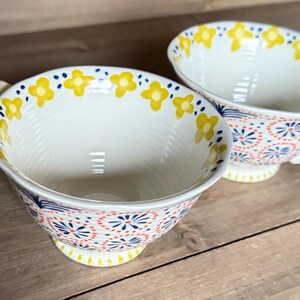 Op de afbeelding: Twee witte keramische theekopjes met oren, versierd met gele bloempatronen rond de randen en kleurrijke bloemen- en geometrische ontwerpen aan de buitenkant. De kopjes staan op een houten oppervlak.