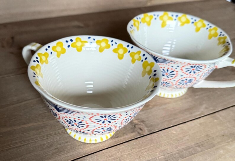 Op de afbeelding: Twee witte keramische theekopjes met oren, versierd met gele bloempatronen rond de randen en kleurrijke bloemen- en geometrische ontwerpen aan de buitenkant. De kopjes staan op een houten oppervlak.