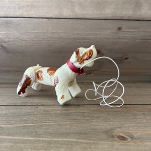 May include: A white and brown plastic toy dog with a red collar and a white string attached to its collar. The dog is in a playful pose with its head tilted back and its tongue sticking out.