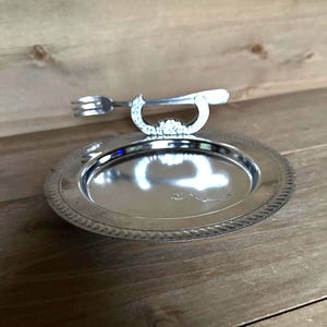 May include: A silver-colored serving tray with a decorative handle and a fork-shaped utensil. The tray has a detailed edge design and sits on a wooden surface. The handle is ornate, adding a touch of elegance.