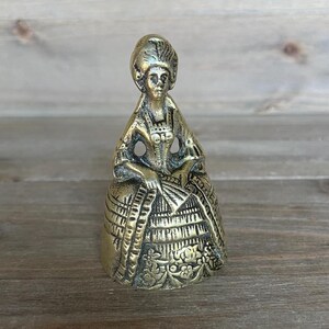Könnte beinhalten: Messingfigur einer Frau in historischer Kleidung, die einen Fächer hält. Die Figur weist detaillierte Kleidung und Haare auf und hat ein leicht gealtertes Aussehen. Die Figur steht auf einer Holzoberfläche.