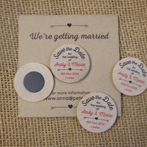 May include: Three round wooden save the date magnets with the text "Save the Date" in a black script font. The text "for the wedding of Andy & Marie" is in a red script font. The date "8th Nov 2016" and the location "London" are in black text.