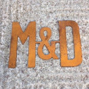 Op de afbeelding: Roestige metalen letters die "M&D" spellen in een decoratief lettertype. De letters zijn bevestigd aan een ruw, grijs oppervlak.