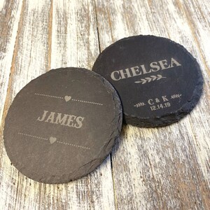 Può includere: Due sottobicchieri rotondi in ardesia nera con nomi incisi. Il sottobicchiere superiore dice "Chelsea" con un design a ramo frondoso e "C & K 12.14.19". Il sottobicchiere inferiore dice "JAMES" con un design a cuore.