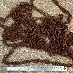 May include: A tangled pile of rusty brown metal chain links. A metal ruler with inch markings is at the bottom of the image, showing the chain's scale. The chain is lying on a rough, gray concrete surface.