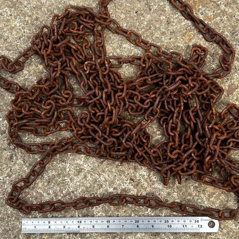 May include: A tangled pile of rusty brown metal chain links. A metal ruler with inch markings is at the bottom of the image, showing the chain's scale. The chain is lying on a rough, gray concrete surface.