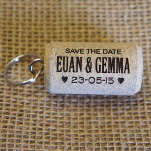 May include: A cork keychain with a black inscription that reads "Save the Date Euan & Gemma 23-05-15" with a heart symbol on either side of the date.
