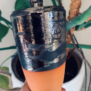 Op de afbeelding: Een keramische fles met een zwarte en zilveren glazuur. De fles heeft een ronde bovenkant en een smalle hals. De bodem van de fles is terracottakleurig.