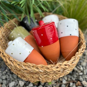 Op de afbeelding: Een rieten mand gevuld met verschillende terracotta-kleurige keramische potten. De potten hebben een cilindrische bovenkant in verschillende kleuren, waaronder rood, wit, zwart en groen, elk versierd met witte stippen. De mand staat op een bed van kleine grijze stenen.