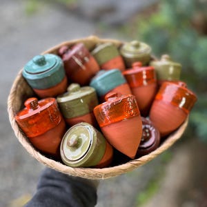 Op de afbeelding: Een gevlochten mand vol kleine, eikelvormige keramische potten. De potten zijn in verschillende kleuren, waaronder oranje, groen en blauwgroen, met bijpassende deksels. De potten hebben een glanzende afwerking.