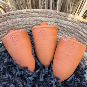 Peut inclure: Trois bouchons de bouteille en céramique de couleur terre cuite avec un motif circulaire en relief sur le dessus. Les bouchons sont placés dans un panier rempli de papier déchiqueté noir.