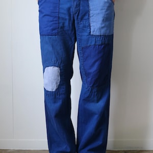 Puede incluir: Pantalones azules con parches de tela en varios tonos de azul cosidos en las piernas y los bolsillos. Los pantalones tienen costuras visibles de hilo oscuro alrededor de los bordes de los parches, creando una apariencia texturizada al frente.
