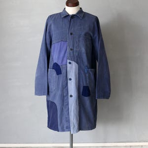 Puede incluir: Una chaqueta de trabajo larga de mezclilla azul con detalles de patchwork. La chaqueta tiene un cierre de botones en la parte delantera y mangas largas. Se muestra en un maniquí.