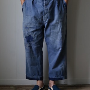 May include: Blue work pants with patches and visible stitching. The pants have a wide leg and are worn with suspenders and blue slip-on shoes. The pants appear to be made of a durable fabric.