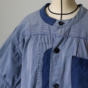 Può includere: Una camicia a righe blu e bianche con maniche corte, colletto rotondo e chiusura con bottoni. La camicia presenta toppe e una tasca blu scuro, con cuciture decorative.