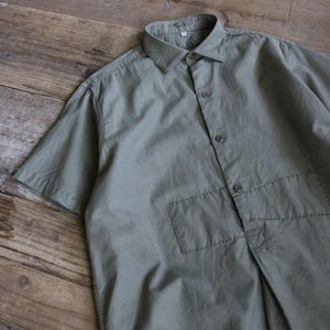 以下が含まれることがあります： ボタン留めのフロントと胸ポケットが付いた、オリーブグリーンの半袖シャツ。 シャツはクラシックな襟で、軽量の生地で作られています。 シャツは木製の表面に平らに置かれています。