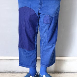 以下が含まれることがあります： 複数のパッチとステッチのディテールが施された青いデニムパンツ。パンツは足首でカフされており、青い靴下と青いキャンバスのスリップオンシューズを履いています。