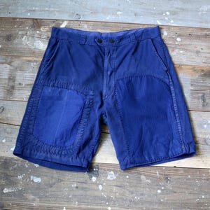 May include: Indigo blue shorts with large, reinforced patches on the front. The shorts feature a button closure and pockets. The garment is made of a robust fabric, possibly cotton or a similar material.