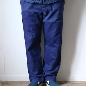 May include: Deep blue trousers featuring reinforced knee patches and visible vertical stitching details along the legs. The trousers have a straight-leg cut and are paired with blue slip-on shoes with white rubber soles, creating a uniform look.