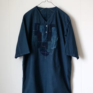 May include: A dark blue, short-sleeved shirt with a V-neck and three buttons. The shirt features a patchwork design on the front, with visible white stitching. The shirt is hanging on a metal hanger against a white wall.