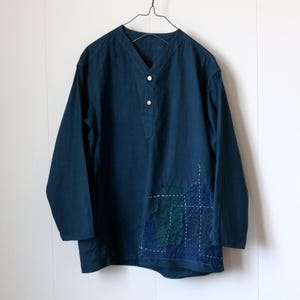 May include: A long-sleeved, navy blue shirt with a V-neck and three white buttons. The shirt features a patchwork design on the lower right side, with visible white stitching. The shirt is hanging on a metal hanger.