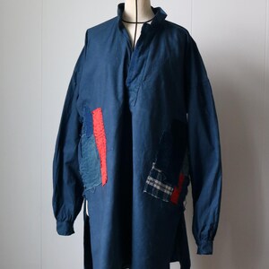 以下が含まれることがあります： マネキンに展示された、長袖でスタンドカラーのネイビーブルーのチュニック。赤、青、チェック柄の生地のパッチが付いています。ゆったりとしたフィット感で、柔らかい素材で作られています。