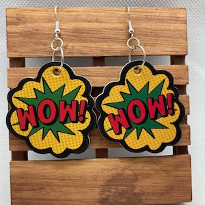 May include: A pair of comic book style earrings. Each earring features a yellow and black cloud shape with the word "WOW!" in red letters on a green burst. The earrings hang from silver-colored hooks.