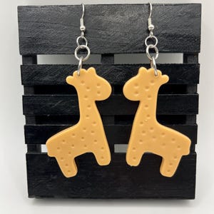 Könnte beinhalten: Ein Paar hellorangefarbene Ohrringe in Form einer Giraffe mit kleinen braunen Punkten. Die Ohrringe hängen an silberfarbenen Fischhaken-Ohrdrähten. Die Giraffen werden auf einer schwarzen Holzpalette ausgestellt.