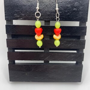 Könnte beinhalten: Hängeohrringe mit Frucht-Design. Jeder Ohrring hat einen silbernen Haken, grüne Perlen, rote herzförmige Perlen, Orangenscheiben und kleine silberne Perlen. Die Ohrringe werden auf einer schwarzen Holzoberfläche präsentiert.