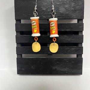 May include: Novelty earrings with miniature Pringles cans, displaying the "Pringles Original" logo in orange and white, with a golden chip pendant. The earrings are suspended from silver-coloured fish hook ear wires.