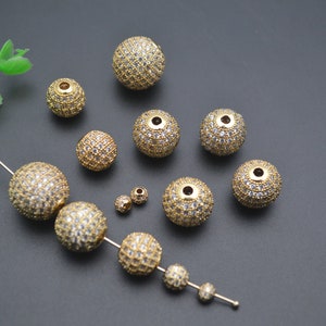 May include: A collection of gold-toned round beads with clear rhinestones. The beads are of various sizes and are scattered on a gray surface.