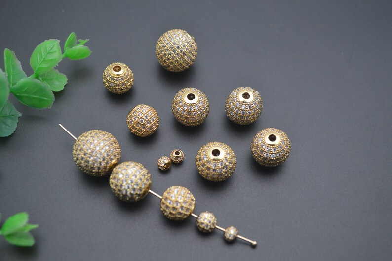 May include: A collection of gold-toned round beads with clear rhinestones. The beads are of various sizes and are scattered on a gray surface.