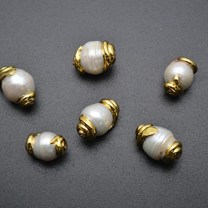 May include: Six white oval pearl beads with gold accents. The beads are arranged in a semi-circle on a dark grey surface.