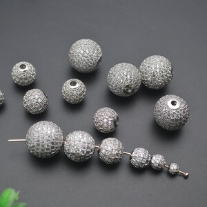 May include: A collection of silver round beads with a sparkly finish. The beads are various sizes and are scattered on a gray surface. Some of the beads are strung together on a silver wire.