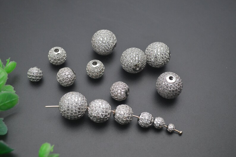 May include: A collection of silver round beads with a sparkly finish. The beads are various sizes and are scattered on a gray surface. Some of the beads are strung together on a silver wire.