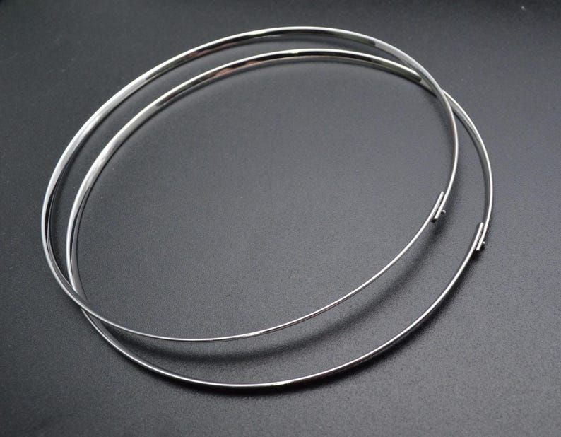 May include: Two silver-colored, thin metal necklaces or chokers, each forming a complete circle. The necklaces are slightly overlapping, with a simple clasp visible on each. The background is a solid, dark gray.