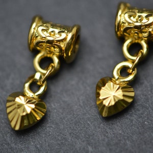 Puede incluir: Dos colgantes de coraz&oacute;n de metal dorado con dise&ntilde;os intrincados. Cada colgante est&aacute; unido a una peque&ntilde;a cadena dorada y una cuenta decorativa en forma de barril.