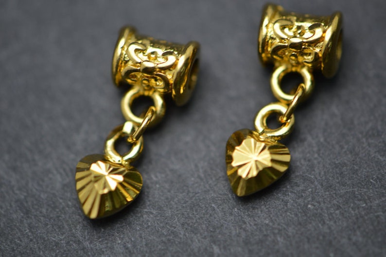 Puede incluir: Dos colgantes de coraz&oacute;n de metal dorado con dise&ntilde;os intrincados. Cada colgante est&aacute; unido a una peque&ntilde;a cadena dorada y una cuenta decorativa en forma de barril.