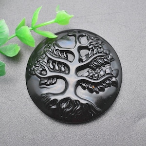 May include: Black obsidian pendant carved with a detailed tree of life design. The pendant is round and has a smooth, polished finish.
