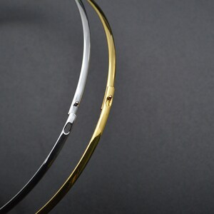 May include: Two metal necklaces, one silver and one gold, are displayed against a dark gray background. The necklaces are thin, circular, and have a clasp closure. The gold necklace is a bright, shiny color.