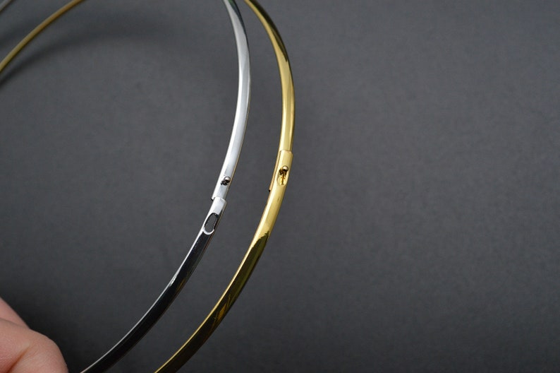 May include: Two metal necklaces, one silver and one gold, are displayed against a dark gray background. The necklaces are thin, circular, and have a clasp closure. The gold necklace is a bright, shiny color.