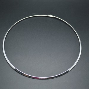May include: A silver-colored choker necklace with a simple, minimalist design. The necklace is a rigid, circular band with a clasp closure. The jewelry is displayed against a dark background, highlighting its reflective surface.