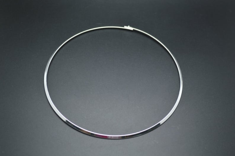 May include: A silver-colored choker necklace with a simple, minimalist design. The necklace is a rigid, circular band with a clasp closure. The jewelry is displayed against a dark background, highlighting its reflective surface.