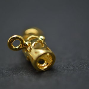 May include: Close-up of a gold-colored jewelry component with a cylindrical shape and intricate cut-out designs. The component has a small loop at one end and a hole at the other. The surface reflects light, highlighting the details.