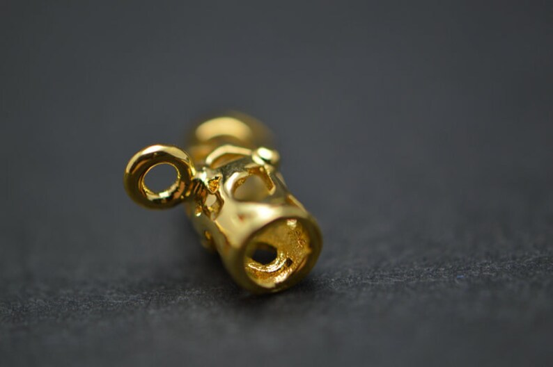 May include: Close-up of a gold-colored jewelry component with a cylindrical shape and intricate cut-out designs. The component has a small loop at one end and a hole at the other. The surface reflects light, highlighting the details.