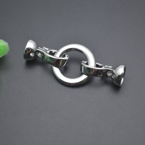 May include: A polished, silver-coloured clasp featuring a central circular ring. The clasp has two rounded ends with a hinged mechanism for opening and closing. Likely used for jewellery or accessories.